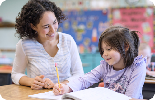 Una persona adulta con cabello rizado y una persona infantil con cabello recogido están sentadas en un aula mientras la persona infantil escribe en un cuaderno con un lápiz.

Descripción generada con IA